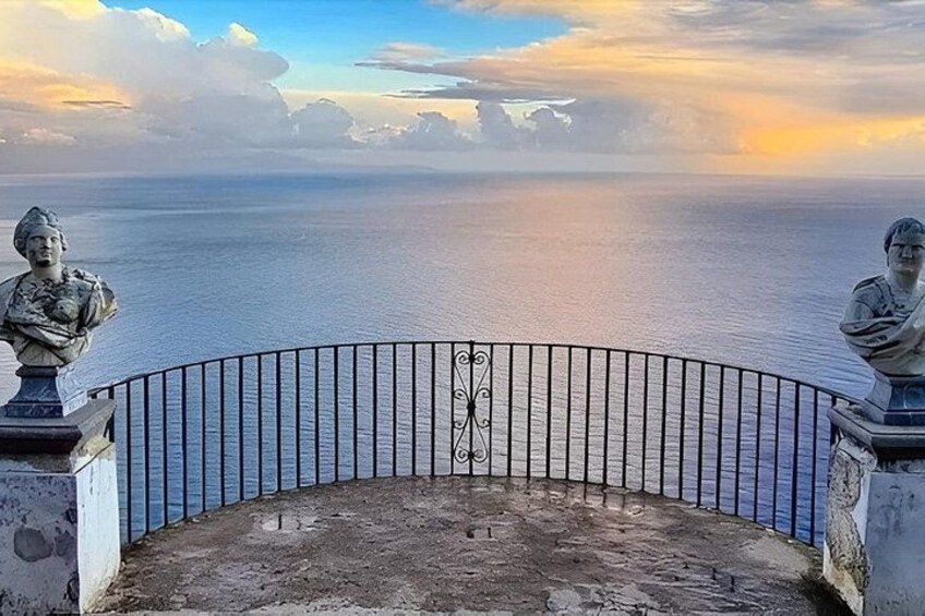A coastal view with a railing, two statues, and a sunset over the sea. 5