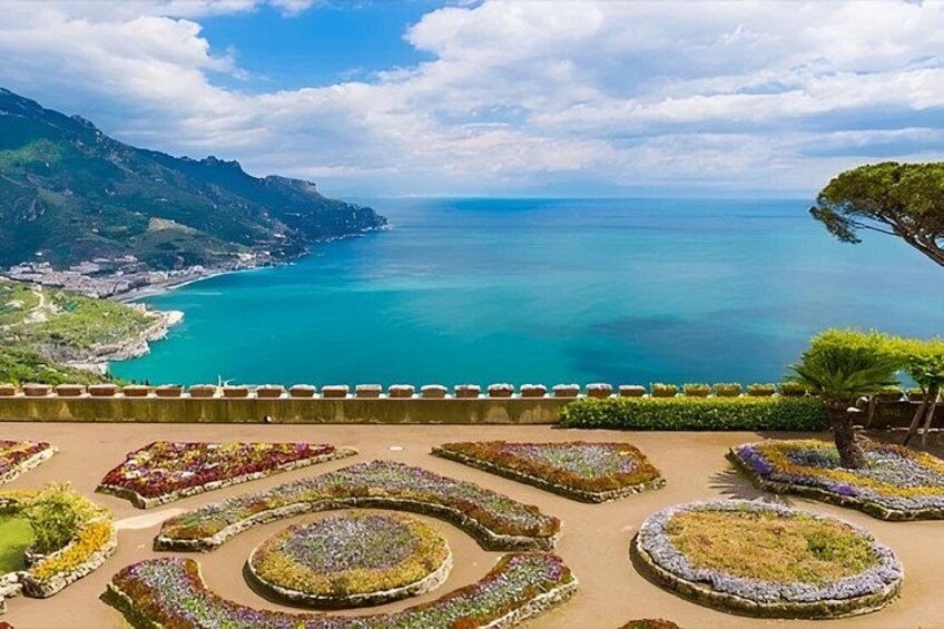 A coastal landscape with a view of the sea, a garden with circular flower beds, and a distant city. 4