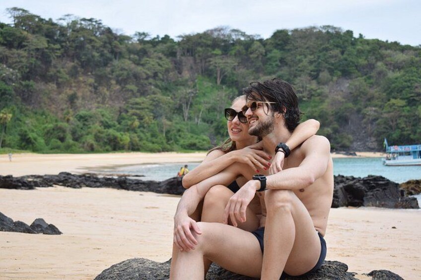 A couple sitting on a rock by a beach with a forested hill in the background. 4