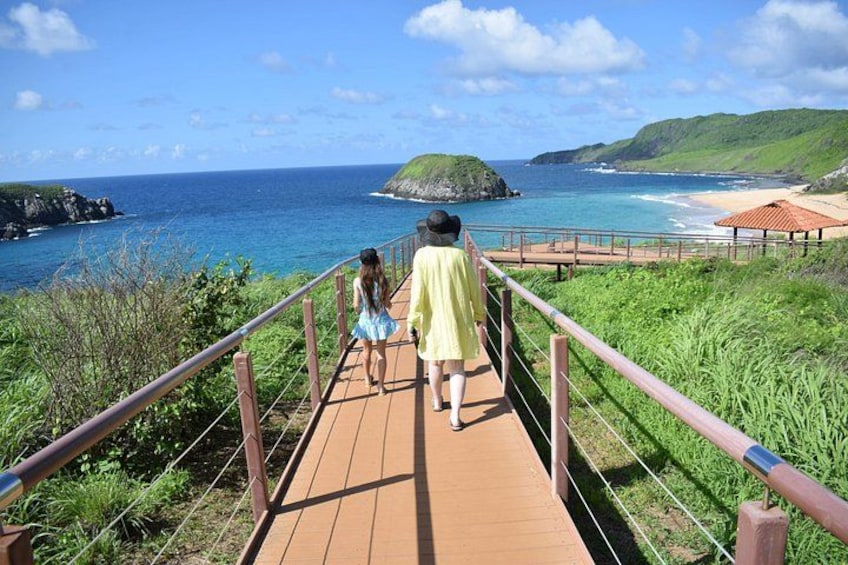 A wooden boardwalk with metal railings, leading to a beach with clear blue water and green hills in the background. 2
