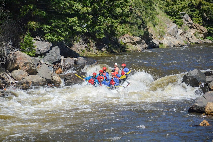 A group of people in blue jackets and red helmets navigating a white-water rafting course with rapids and rocks. 5