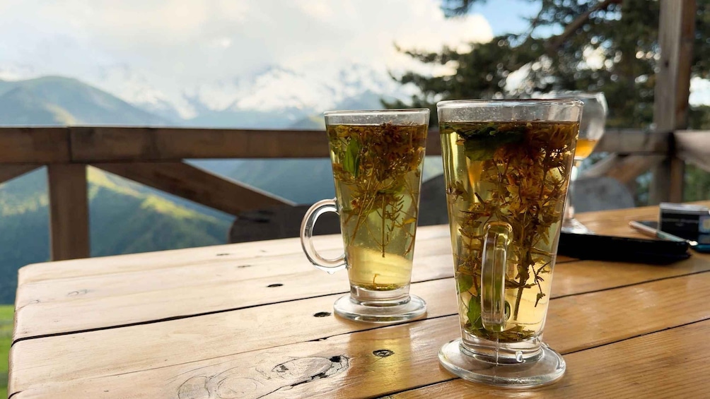 Two clear glasses of tea with herbs on a wooden table with a mountain view in the background. 5