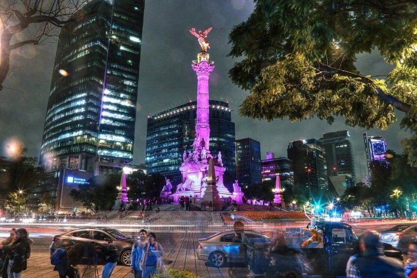 A brightly lit monument with a statue at the top, surrounded by modern buildings and trees. 2