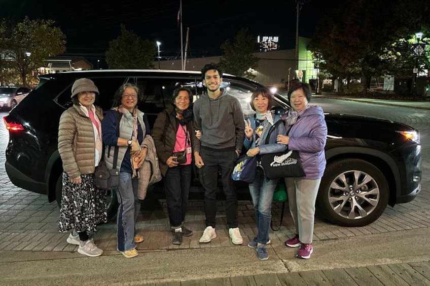 A group of people standing in front of a black car on a paved area at night. 3