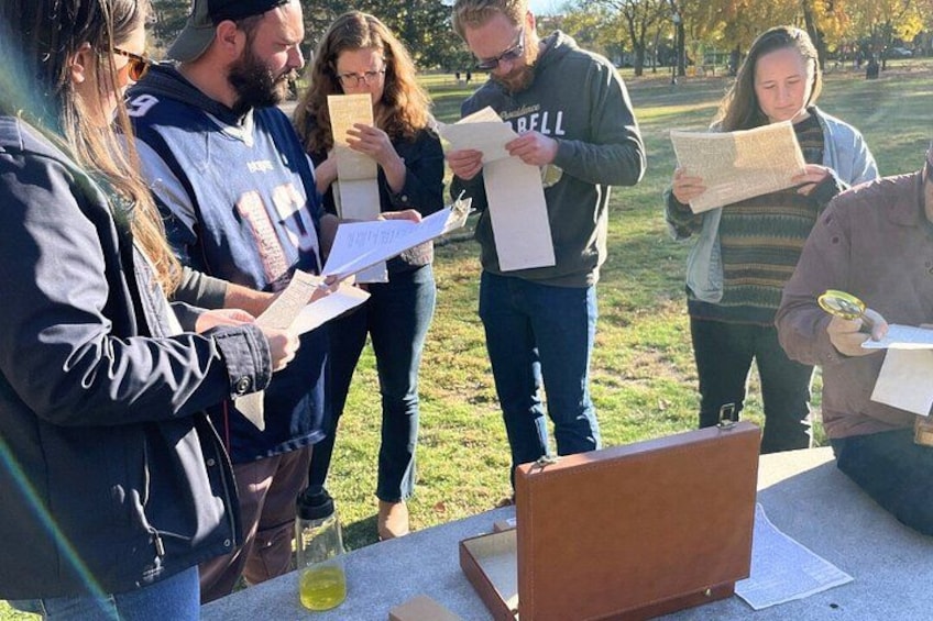 A group of people standing around a table with papers, one person holding a magnifying glass, and a brown leather bag on the table. 2