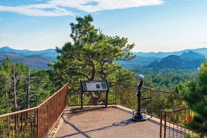 A paved viewing platform with a metal railing, a signpost, and a tree in a mountainous landscape. 4