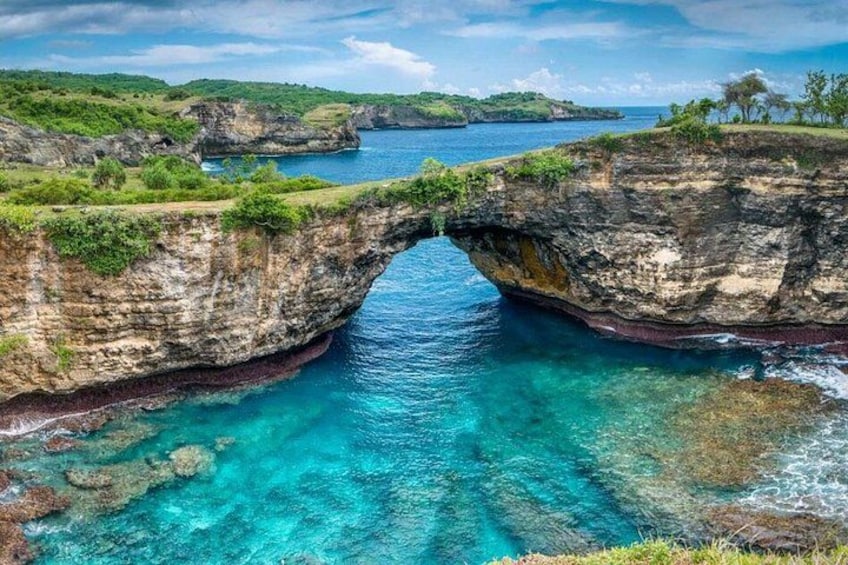 A natural rock formation arching over a clear blue sea, surrounded by lush greenery and distant islands. 1