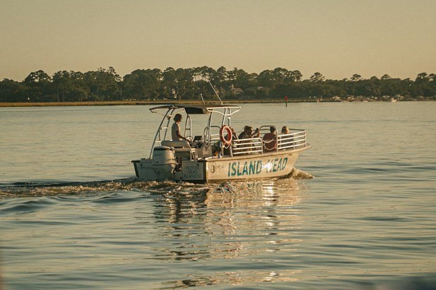 Een boot met de naam 'Island Head' op kalm water, met een bebost strand in de achtergrond. 1