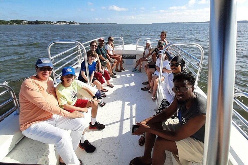 Een groep mensen op een boot met metalen leuningen, zittend en staand, met water en een verre kustlijn op de achtergrond. 4