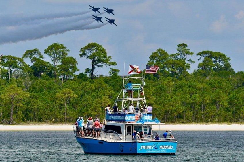 A boat with people on it, a flag, and a jet formation in the sky. 4