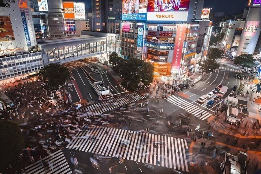 Une intersection urbaine très fréquentée, avec de nombreuses voies de circulation et des passages piétons, entourée d’immeubles commerciaux aux vitrines illuminées. 3