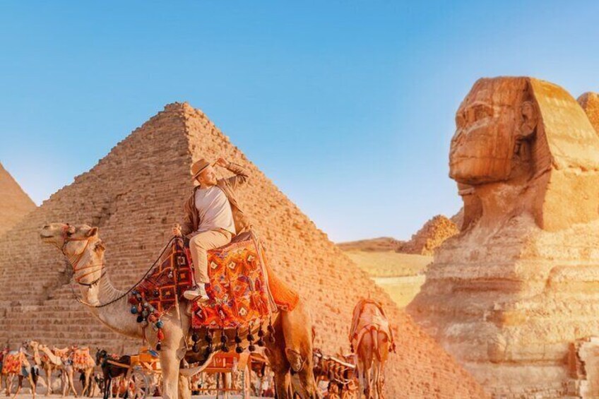 En person som rider på en kamel med en utsmyckad sadel, med en pyramid och den stora sfinxen i bakgrunden. 1