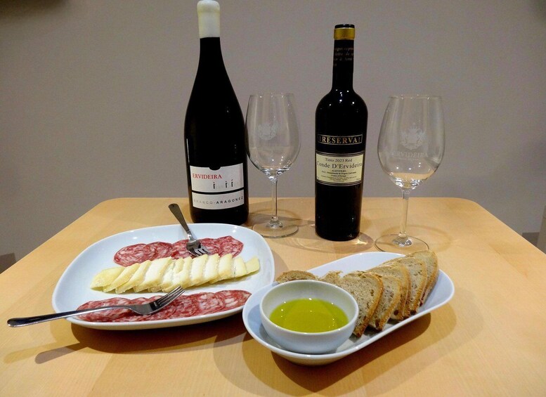 A table with a plate of cured meats, a bowl of olive oil, two wine bottles, and two empty glasses. 1