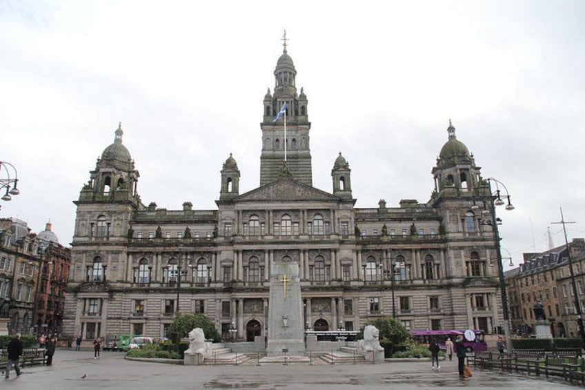 A large historic building with a central tower, a statue in the foreground, and a statue of a person in the background. 3
