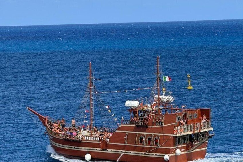 Un barco de madera grande con varios mástiles y banderas, navegando sobre un mar azul. 3