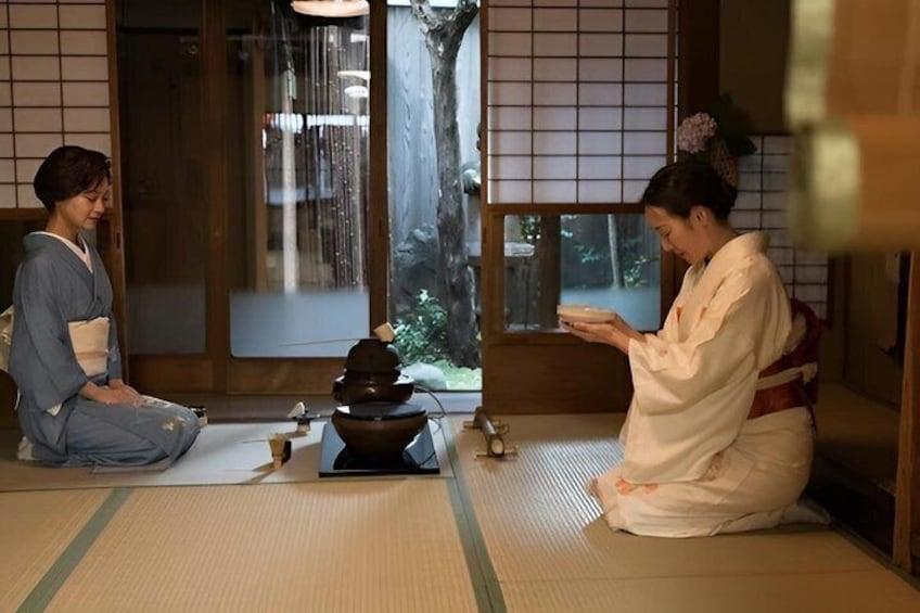 Two people in traditional attire sitting on tatami mats in a room with a wooden floor, a stone basin, and a window with a view of greenery. 1