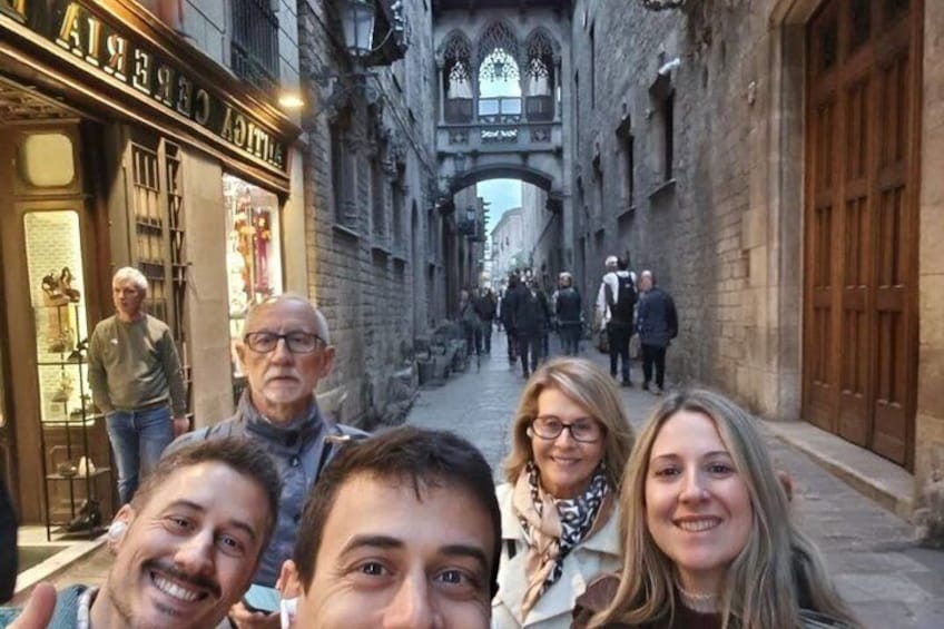 A narrow medieval street with stone buildings, a shop with a sign, and people walking. 1