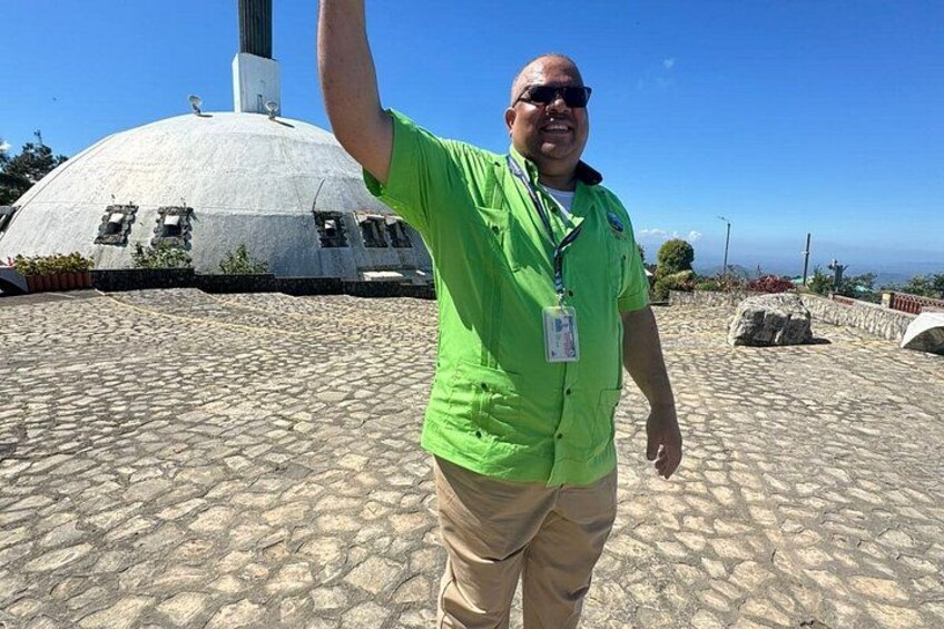 Una persona con camisa verde y pantalones color beige de pie frente a una estructura en forma de cúpula con un asta de bandera. 3