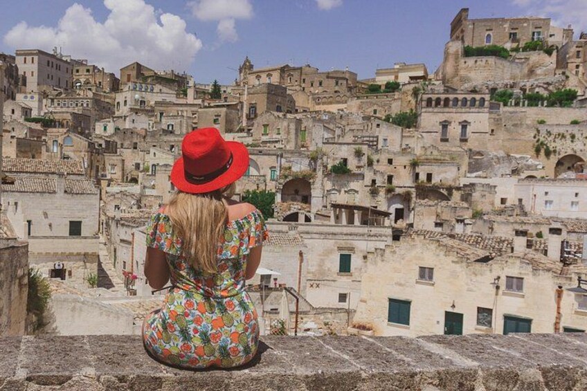 A woman in a red hat and floral dress sitting on a stone ledge overlooking a densely packed historic town with terracotta rooftops. 2