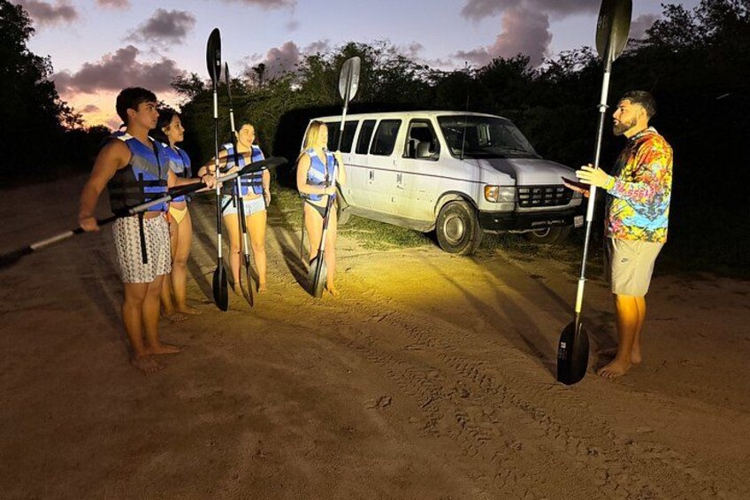 People with kayaks and paddles, a white van, and a sandy area at dusk. 4