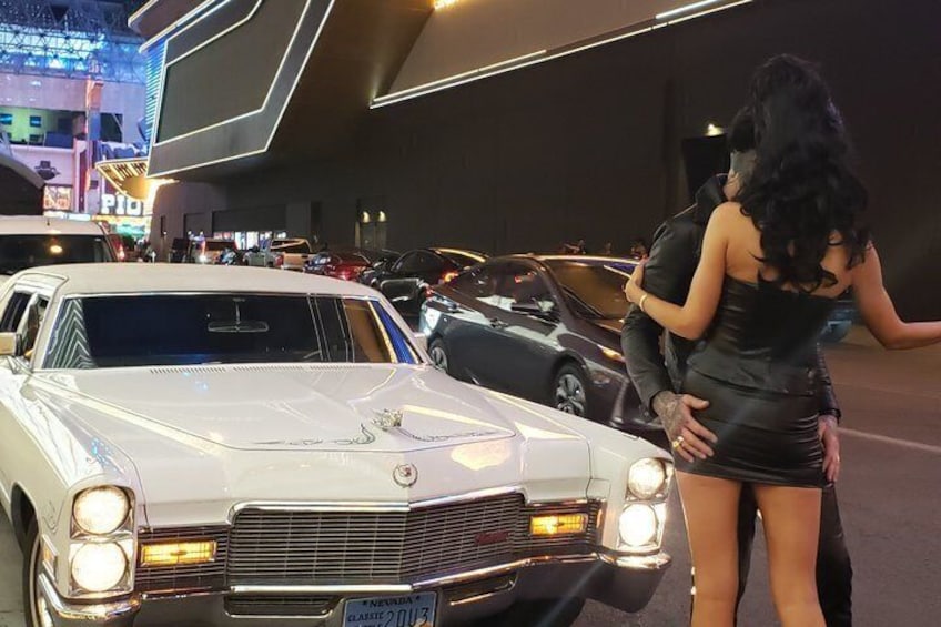 Une voiture ancienne blanche, dotée d’une calandre et de phares bien visibles, est garée dans un espace intérieur bien éclairé, aux côtés d’autres véhicules, avec une enseigne au néon en arrière-plan. 1
