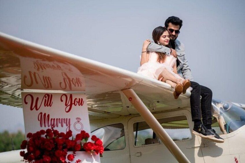 A couple sitting on a small aircraft with a banner that reads 'Will You Marry Me?' 1