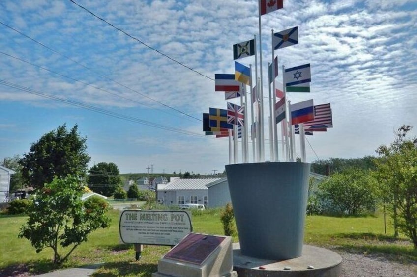 A melting pot sculpture with multiple national flags, surrounded by a sign reading 'THE MELTING POT,' set against a backdrop of a clear sky and a grassy area with trees. 3