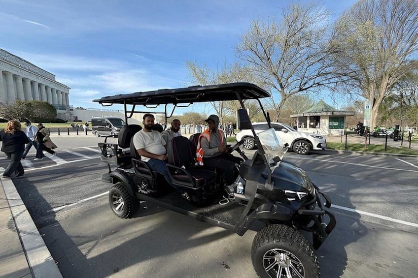 A golf cart with passengers on a paved area with trees and a building in the background. 1