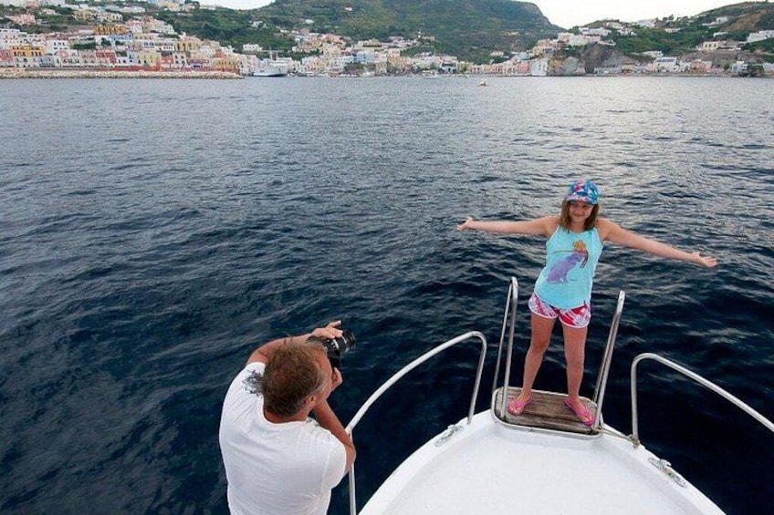 A person standing on a boat's deck with arms outstretched, facing a body of water with a coastal town in the background. 5