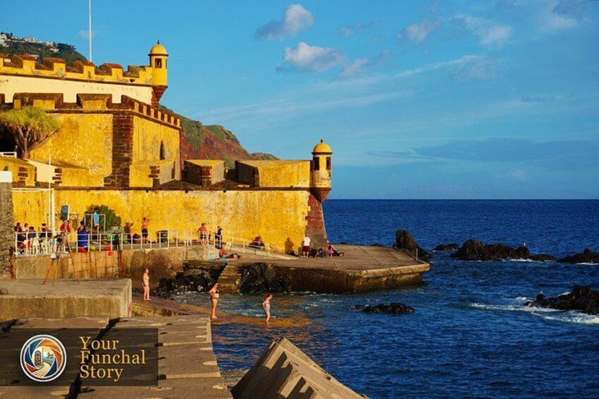 A coastal fort with a lighthouse, people walking along the pier, and the ocean in the background. 1