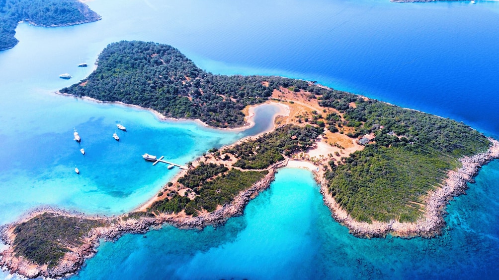 An aerial view of a coastal island with clear turquoise waters, a small dock, and several boats anchored near the shore. 1