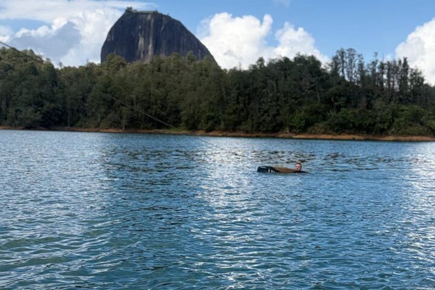 Un lago tranquillo con una persona che nuota, circondato da una fitta foresta e una roccia imponente. 2