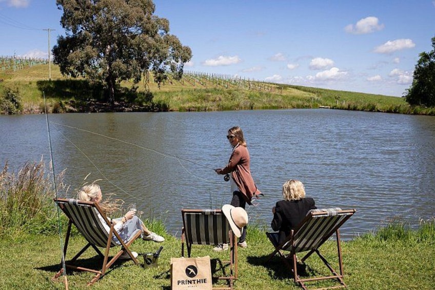 Three people fishing by a lake, with chairs and a sign labeled 'PRINTHE' on the grass. 4