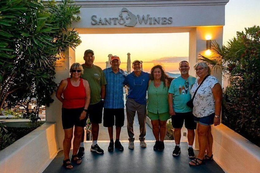En grupp människor som står framför en skylt med texten ”Santo Wines”, med en solnedgång i bakgrunden. 2