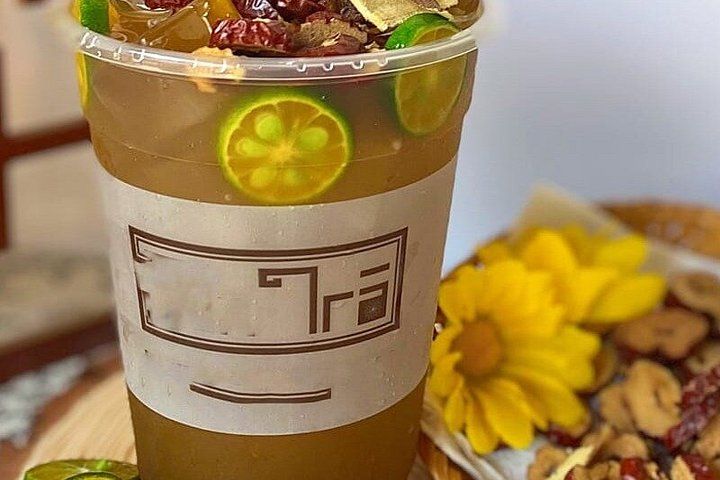 A beverage in a branded cup with a logo, garnished with lime slices and dried fruit, with a blurred background of a table and a flower arrangement. 2