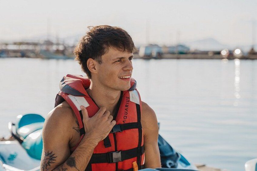 Una persona con chaleco salvavidas rojo con la palabra 'QUADRIUS', de pie junto a una masa de agua con barcos al fondo. 3