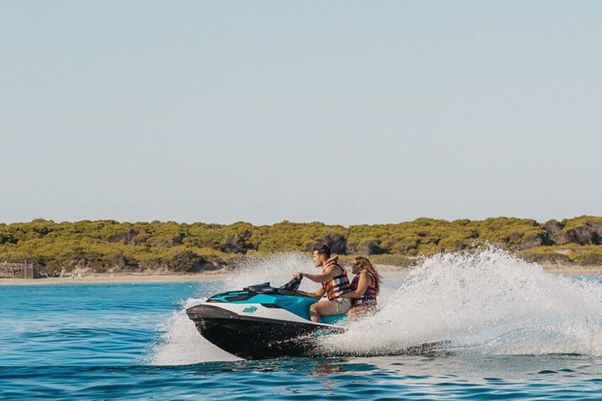 Un jet ski con dos personas avanzando entre el agua, con una playa y vegetación al fondo. 5