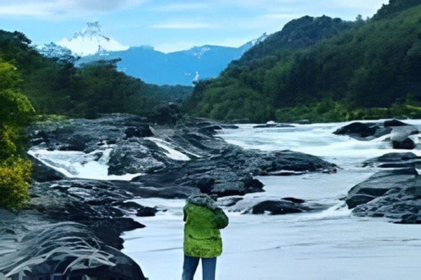 A river with rocks, a person in a green jacket, and mountains in the background. 4