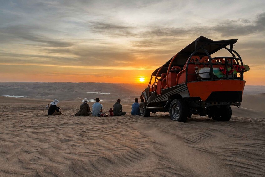 A desert landscape with a group of people sitting on the sand, a dune buggy parked nearby, and the sun setting in the background. 1
