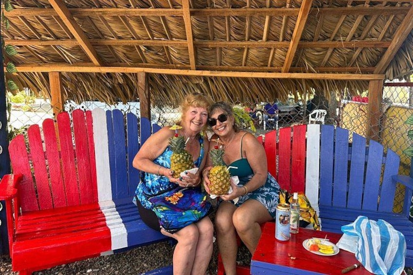 Two people sitting on a bench with a tropical umbrella, holding pineapples, and a table with food and drinks. 2