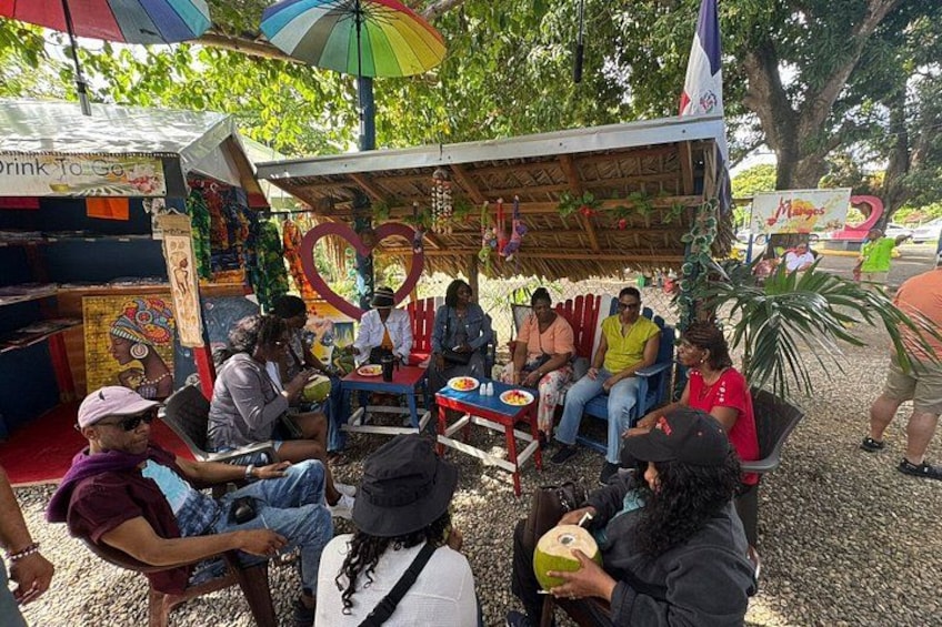 A group of people sitting around a table under a shaded area with a colorful umbrella. 5
