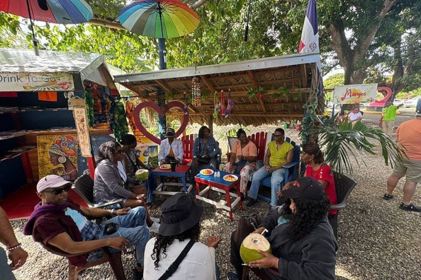 A group of people sitting under a shaded area with a colorful umbrella, surrounded by greenery. 4