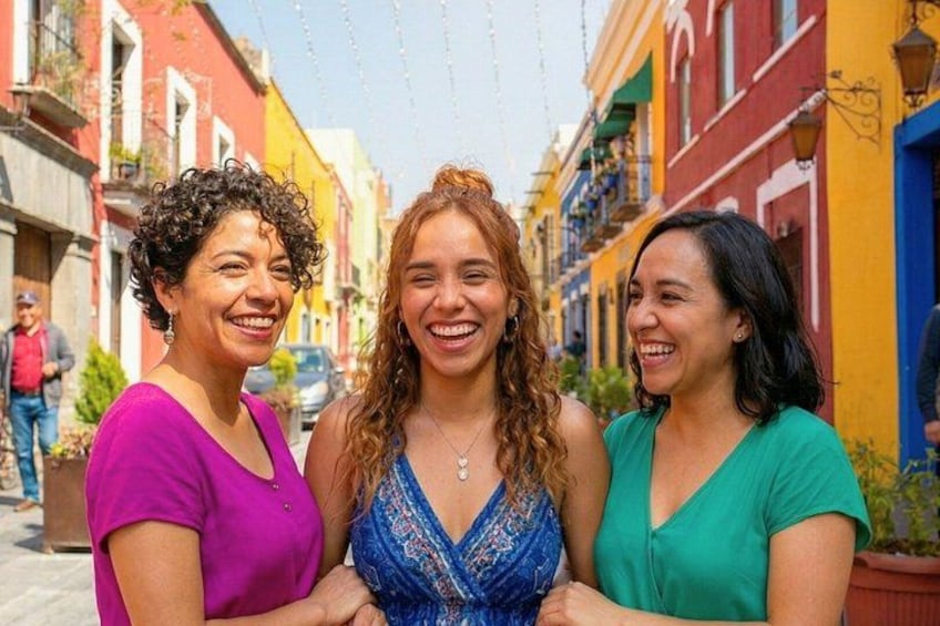 Three women standing on a colorful street with buildings on either side. 1