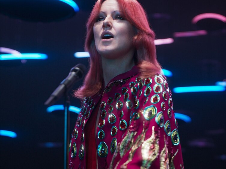 A person with red hair wearing a red and gold patterned outfit, standing in front of a microphone. 4