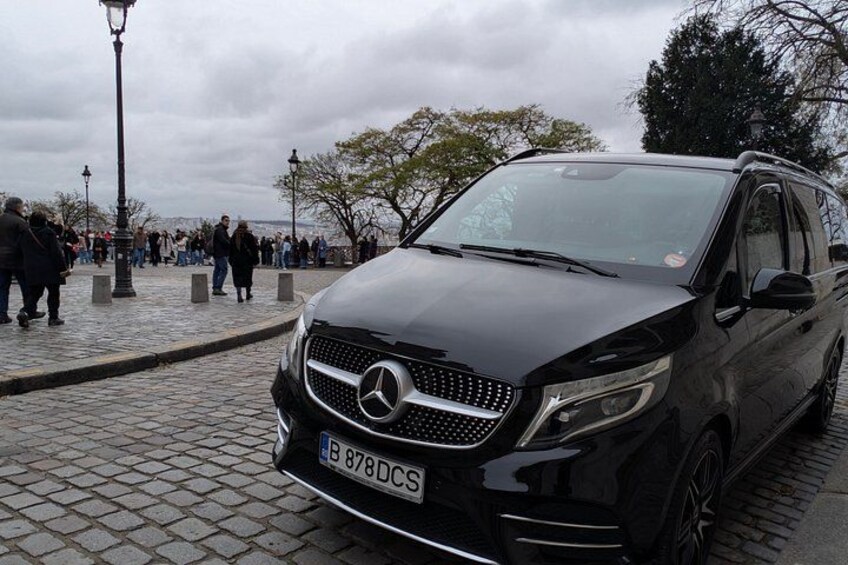 En sort Mercedes-Benz SUV holder parkeret på en brostensbelagt sti, mens fodgængere ses i baggrunden. 1