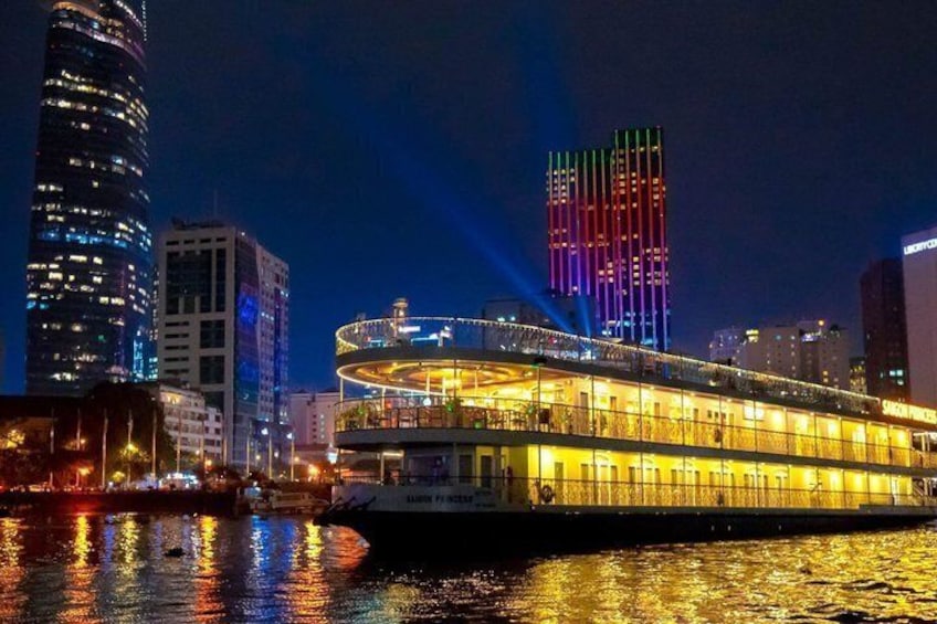 A riverboat with illuminated windows and a deck, docked at night with city buildings in the background. 1