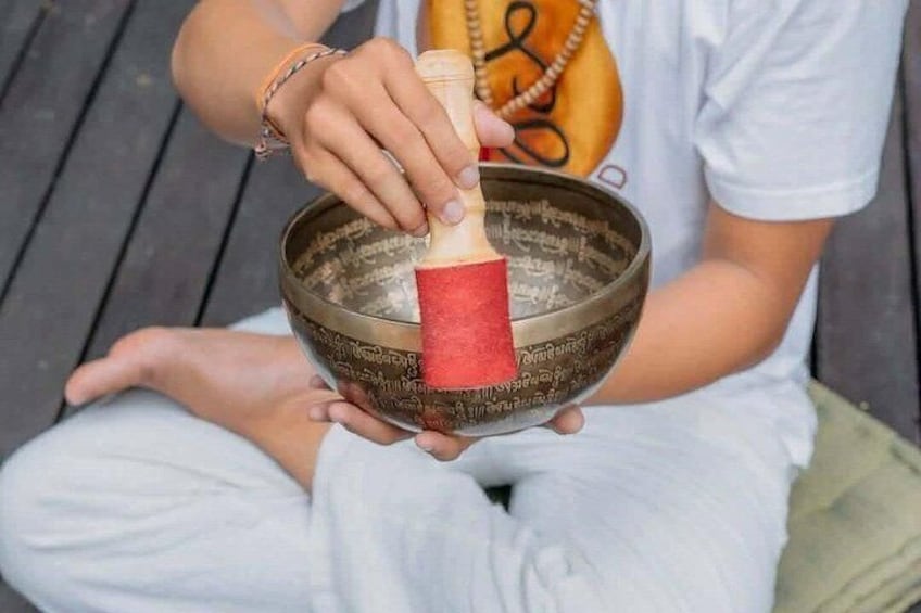 A person sitting on a wooden bench, holding a singing bowl with a red mallet. 2