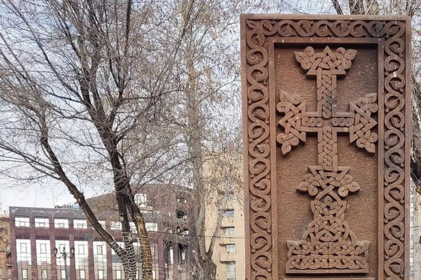 A carved wooden cross with intricate designs, set against a backdrop of leafless trees and buildings. 4