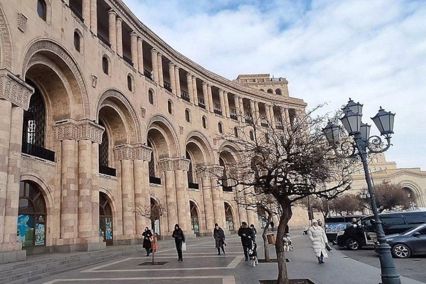 A historic building with arches and columns, a tree, and a street with pedestrians and parked cars. 2