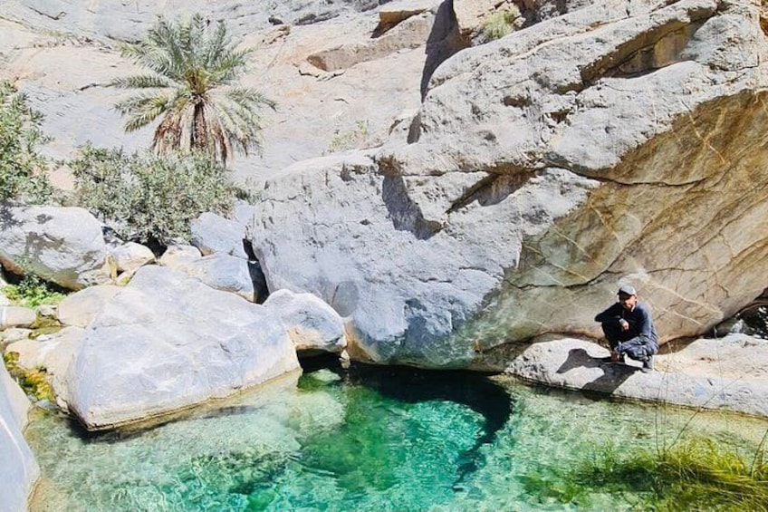 Una persona sentada sobre una roca junto a una piscina de agua cristalina, rodeada de grandes rocas y palmeras. 1
